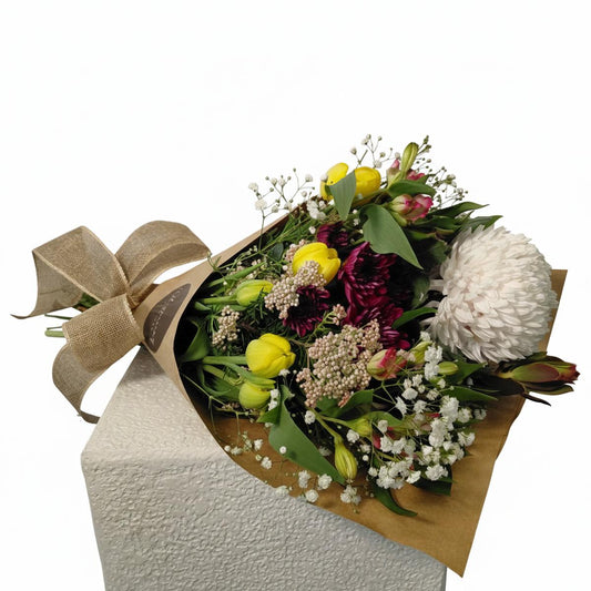 A mixed floral bouquet wrapped in brown kraft paper and tied with a large burlap ribbon, displayed on a textured white pedestal against a white background. The bouquet features yellow tulips, a large white pom-pom flower, burgundy protea, and white baby's breath.