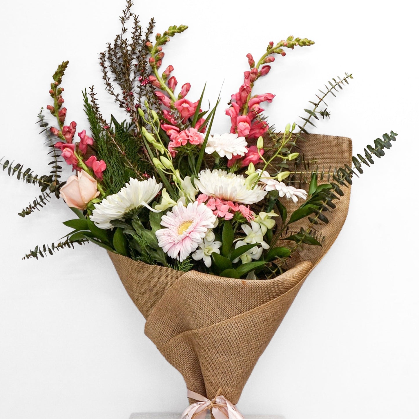 A fresh floral bouquet wrapped in brown burlap paper and tied with a pale pink ribbon, displayed on a white pedestal against a plain white background.