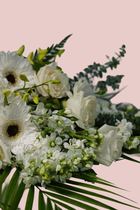 A close-up floral arrangement featuring white gerbera daisies, white roses, small white blossoms, green foliage, and palm leaves against a solid pale pink background.