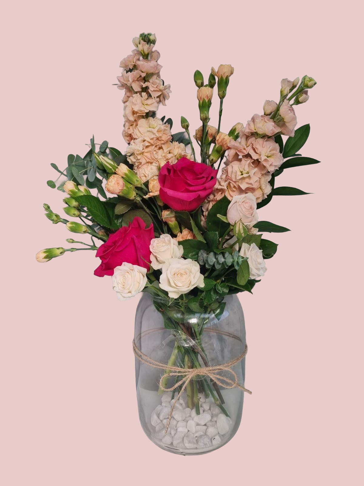 A floral bouquet with pink and white roses, peach carnations, and green foliage arranged in a clear glass jar vase with white stones and twine, on a solid pale pink background.