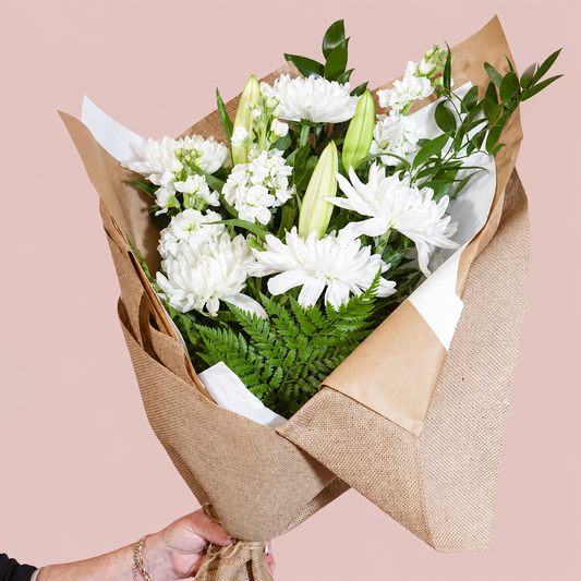A bouquet of white flowers and green foliage wrapped in brown burlap and kraft paper, held by a hand against a solid pink background.