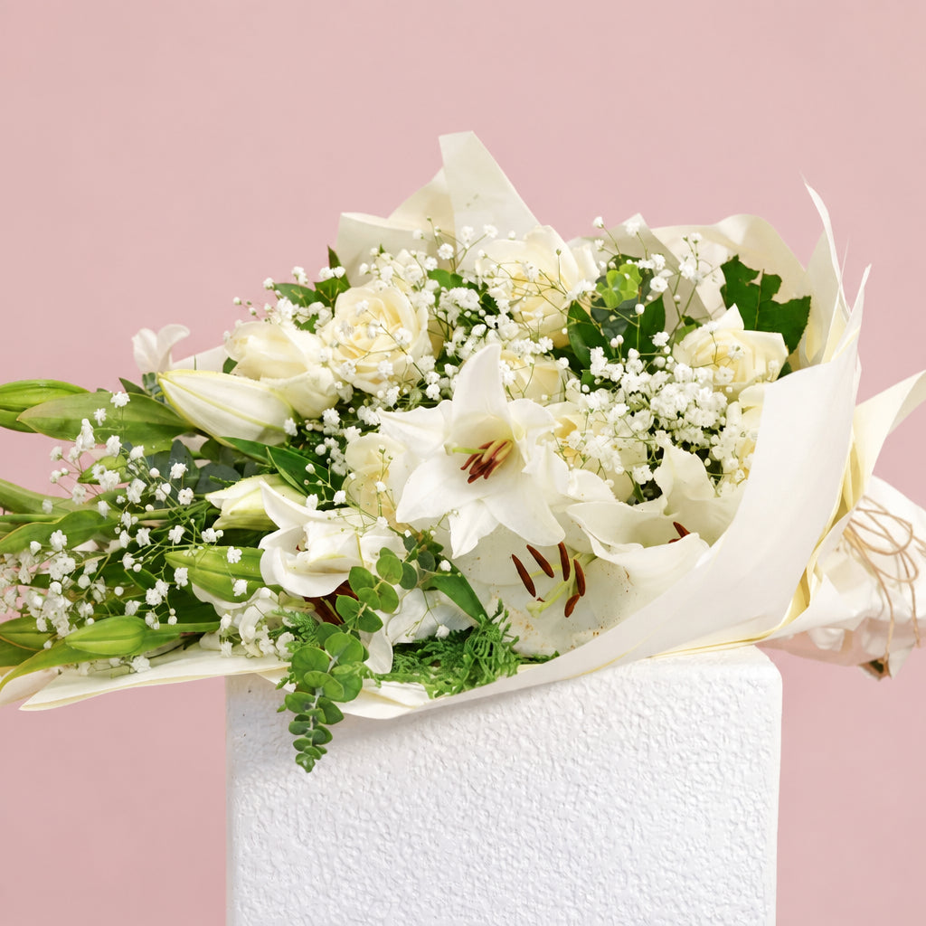 A bouquet of white lilies, white roses, and baby's breath flowers with green leaves, wrapped in white tissue paper and tied with a gold string, displayed on a white pedestal against a solid pink background.