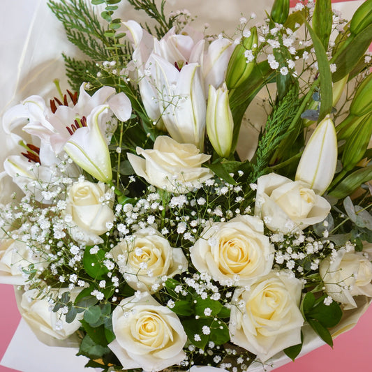 A bouquet of white and cream flowers including lilies, roses, and baby's breath, wrapped in white paper against a solid pink background.