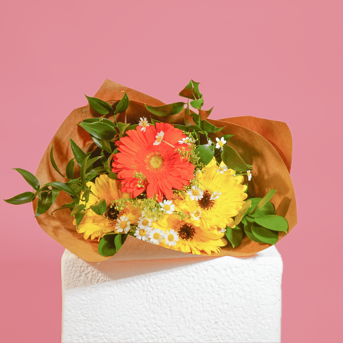 A mixed flower bouquet with a bright red-orange gerbera daisy, yellow gerbera daisies, small white daisies, and green leaves, wrapped in brown paper and displayed on a white pedestal against a solid pink background.