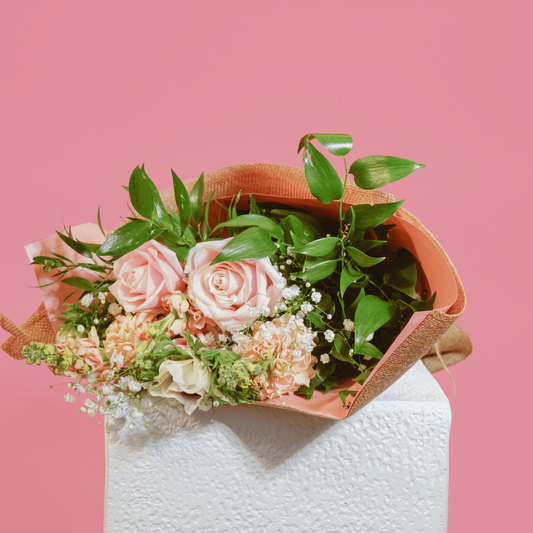 A floral bouquet with pink roses, peach blossoms, and green leaves wrapped in brown paper, displayed on a white pedestal against a solid pink background.