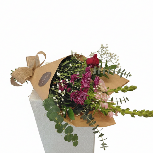 A floral bouquet wrapped in brown kraft paper and tied with a burlap ribbon, displayed on a white pedestal against a white background. The bouquet contains green eucalyptus, pink and purple flowers, and small white filler blooms.