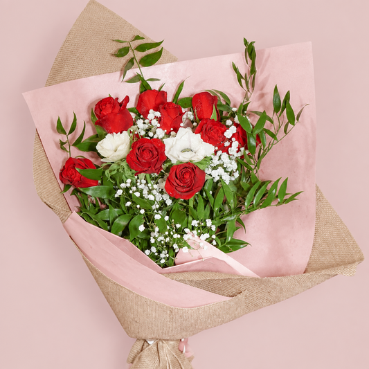 A bouquet of red and white roses with green foliage, wrapped in pink tissue paper and natural burlap, held by a person's hand against a solid pink background.