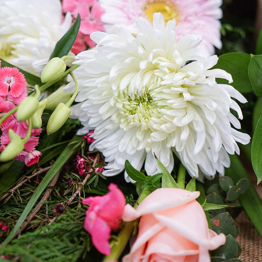 A close-up of a floral arrangement featuring white and light pink flowers, including a large white chrysanthemum, a pale pink rose, pink carnations, and green foliage, set against a burlap-like background.