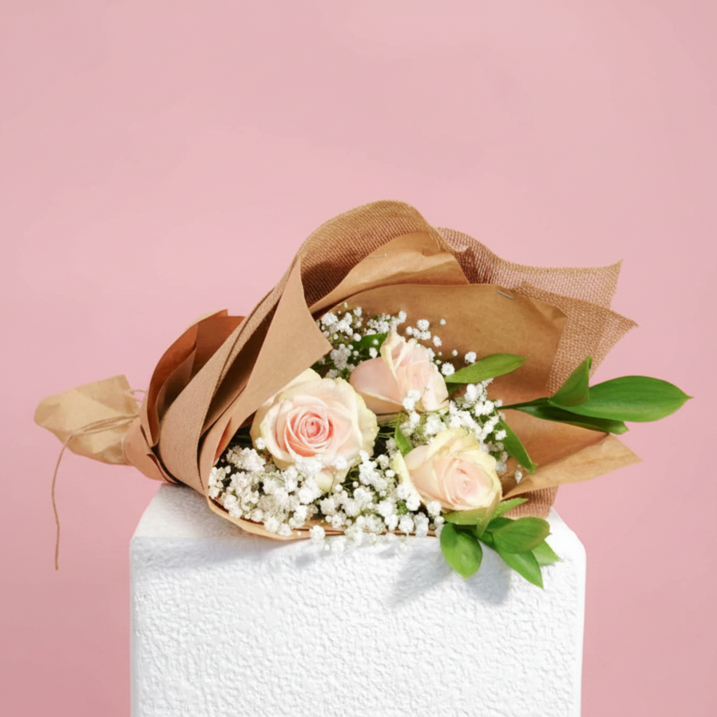 A bouquet of three pale pink roses, white baby's breath, and green leaves, wrapped in brown kraft paper, displayed on a white pedestal against a solid pink background.