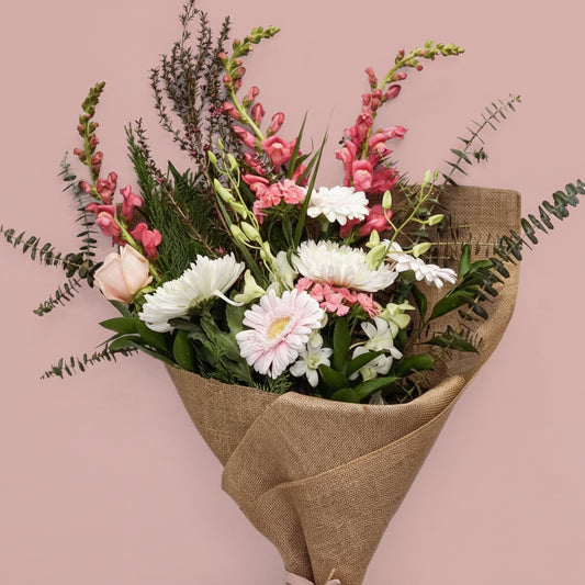 A mixed floral bouquet wrapped in brown burlap paper and tied with a pale pink ribbon, displayed on a white pedestal against a white background.