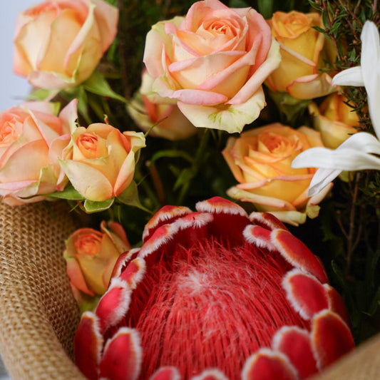 A close-up of a floral arrangement featuring peach and cream roses, pink carnations, white lilies, a large red protea, and green foliage, wrapped in burlap.