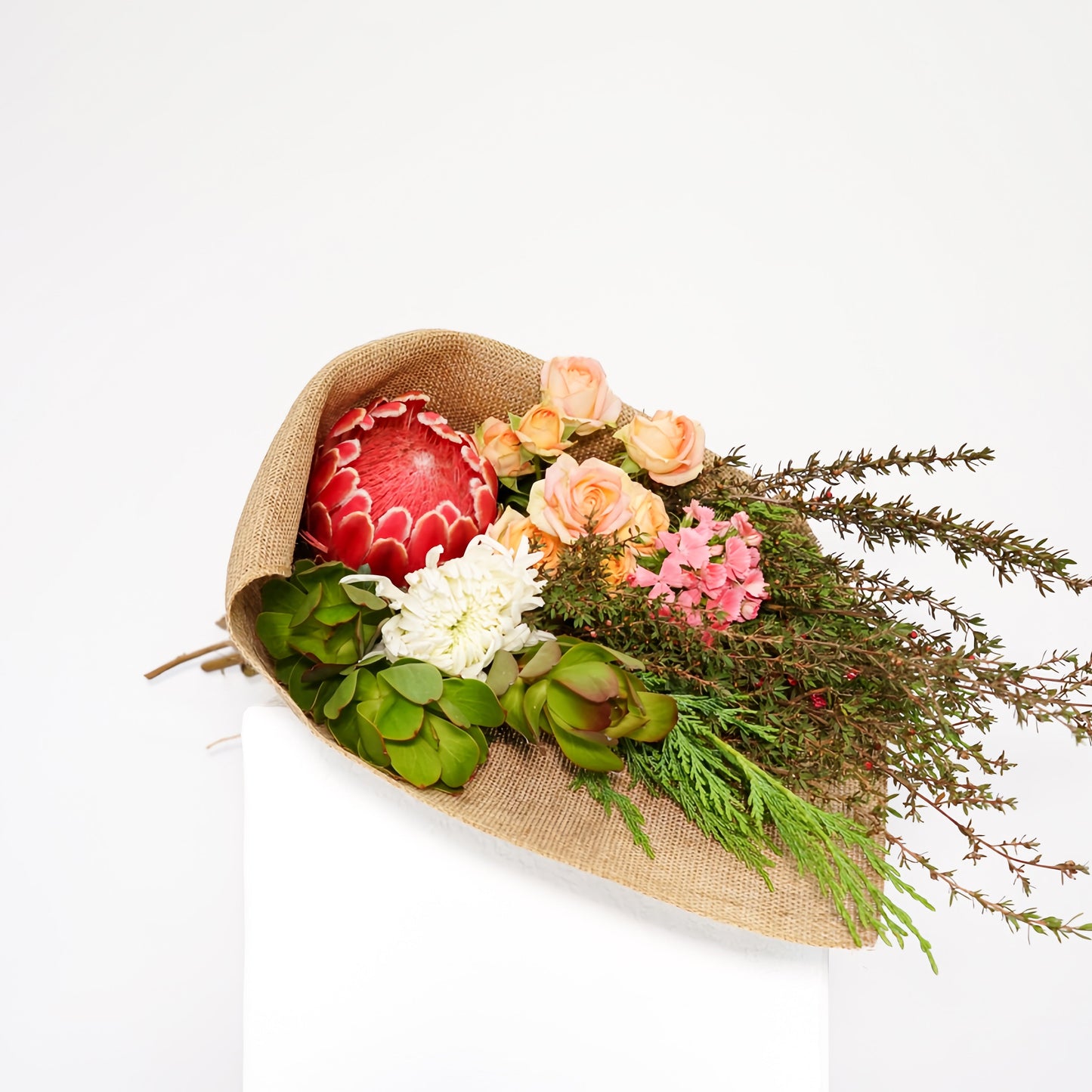 A rustic floral arrangement in a burlap-wrapped paper bag, featuring a red patterned protea, peach and pink roses, white flowers, green foliage, and brown branches, displayed on a white pedestal against a white background.