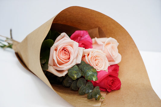 A bouquet of fresh roses wrapped in brown kraft paper, featuring light pink, deep pink, and red roses with green eucalyptus leaves, set against a clean white background.