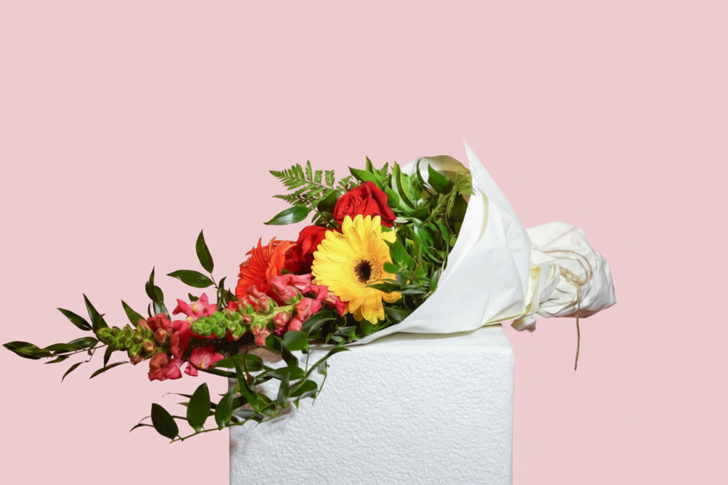 A vibrant floral bouquet wrapped in white paper, featuring a bright yellow gerbera daisy, red roses, pink snapdragons, and green foliage, displayed on a white textured pedestal against a solid pink background.