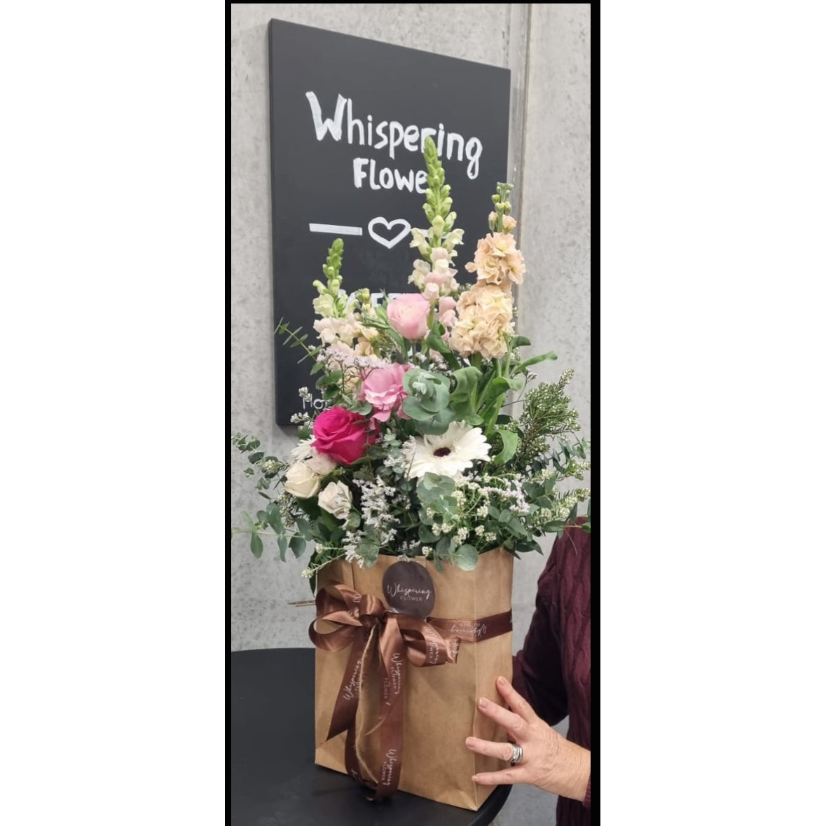 Woman holding a bouquet of flowers with 'Whispering Flowers' sign in the background