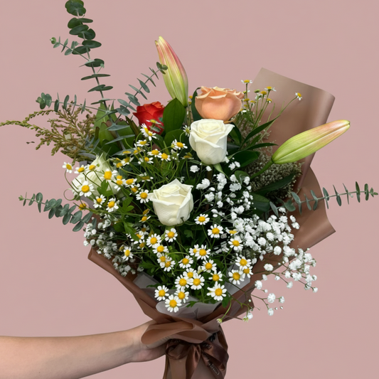 A hand holding a fresh flower bouquet wrapped in brown paper. The bouquet features white and peach roses, lily buds, small white daisies, baby's breath, green eucalyptus stems, and a red rose accent, set against a plain white background.