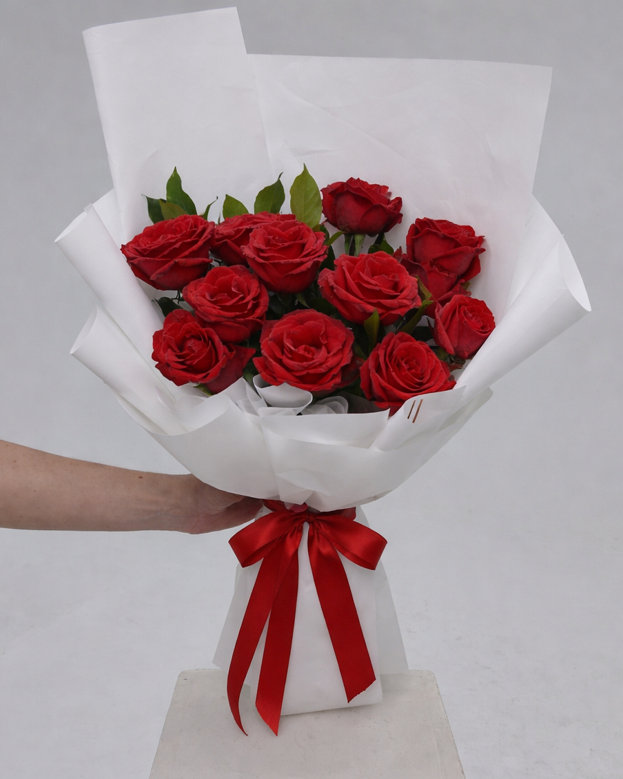 Bouquet of red roses wrapped in white paper with a red ribbon, held by a person on a gray background.