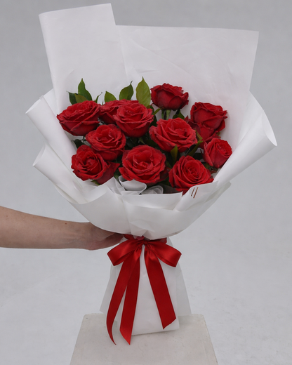 Bouquet of red roses wrapped in white paper with a red ribbon, held by a person on a gray background.