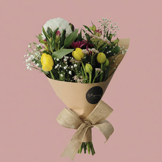 A fresh flower bouquet wrapped in brown kraft paper with a tied ribbon, featuring yellow tulips, white flowers, green foliage, and burgundy accents on a plain white background.
