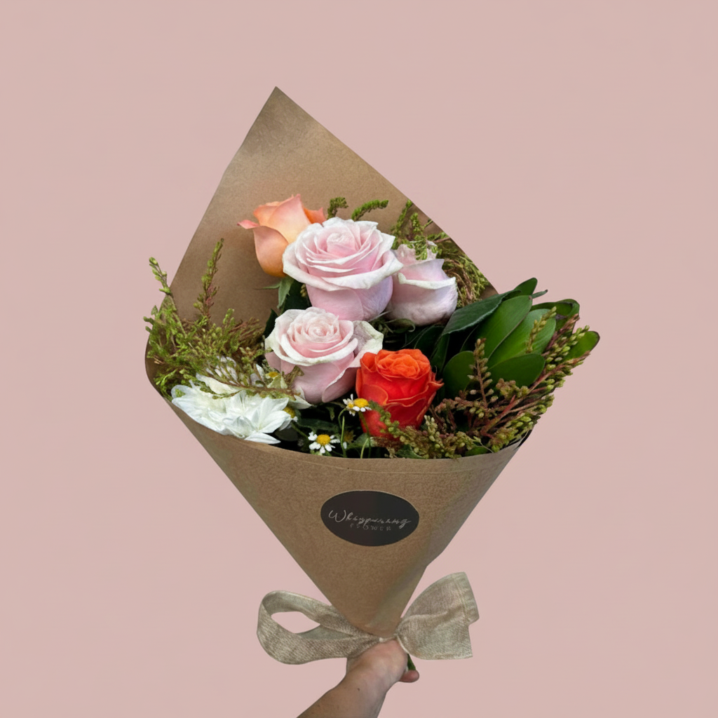 A floral bouquet wrapped in brown kraft paper with a beige ribbon and a small round black label reading 'Wishing Flowers,' held by a hand against a plain white background. The bouquet contains pale pink roses, a bright orange-red rose, white flowers, green foliage, and small yellow accents.
