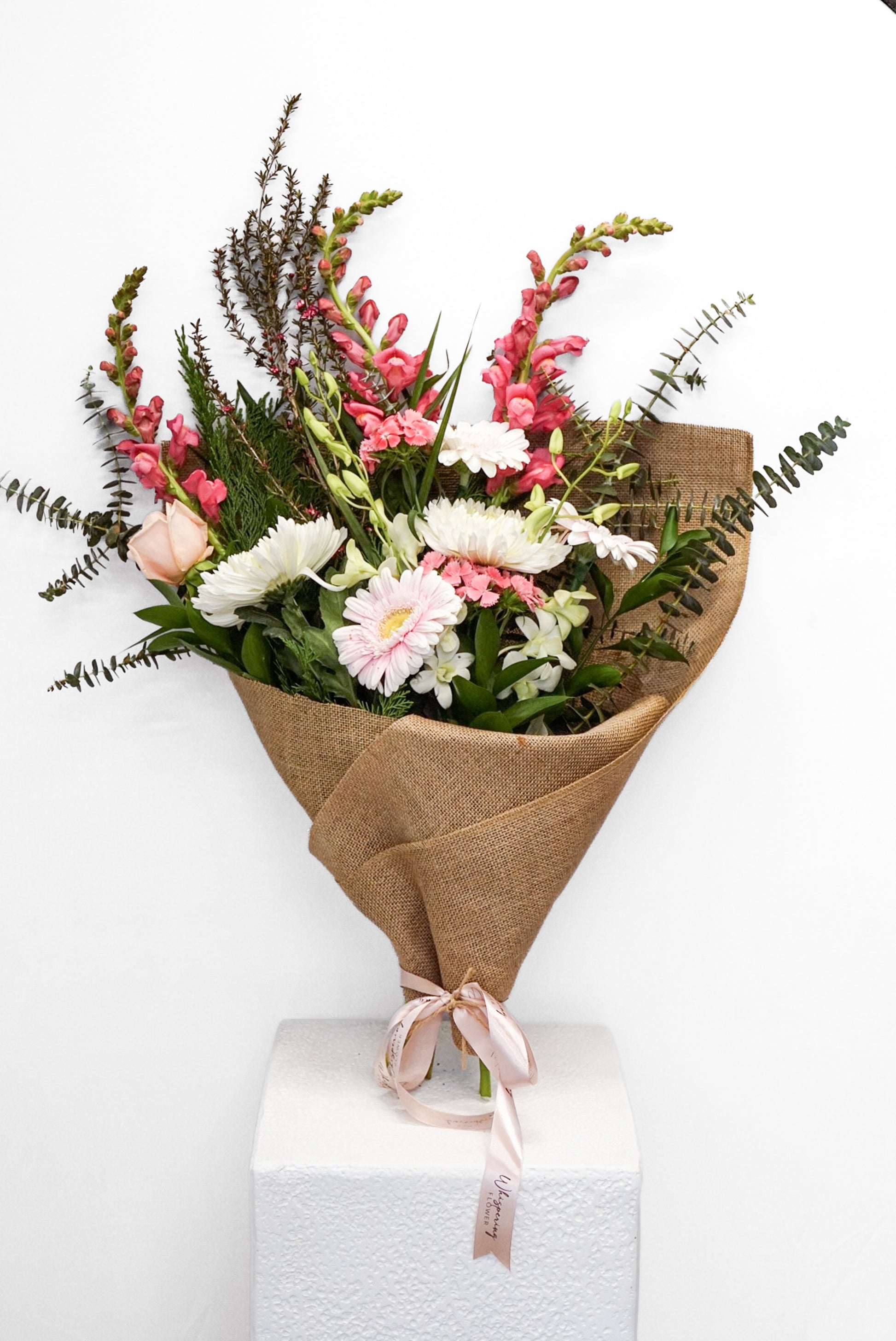 A fresh floral bouquet wrapped in brown burlap paper and tied with a pale pink ribbon, displayed on a white pedestal against a plain white background.
