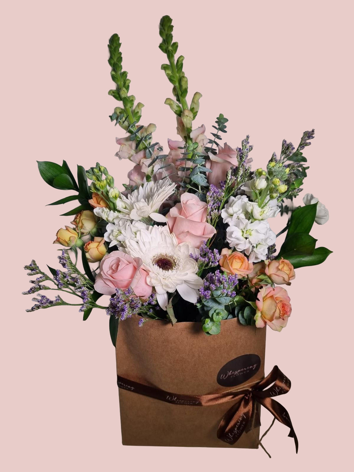 A floral arrangement in a brown kraft paper box with a brown ribbon and bow, featuring pink roses, white daisies, peach geraniums, and green foliage, set against a solid pale pink background.