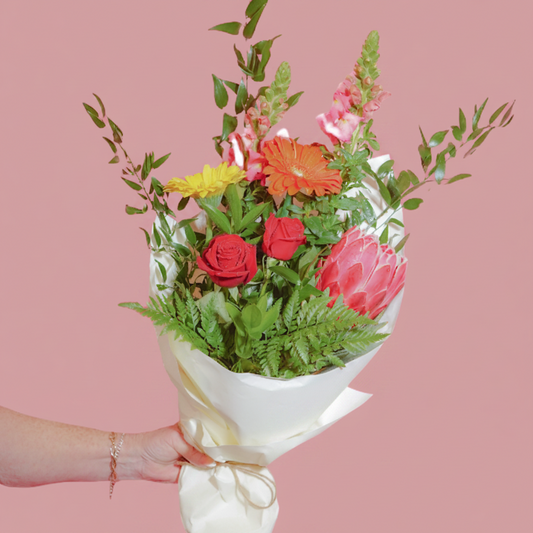 A hand holding a vibrant floral bouquet wrapped in white paper, featuring red roses, a pink protea, orange gerbera daisies, yellow flowers, and green foliage on a solid white background.