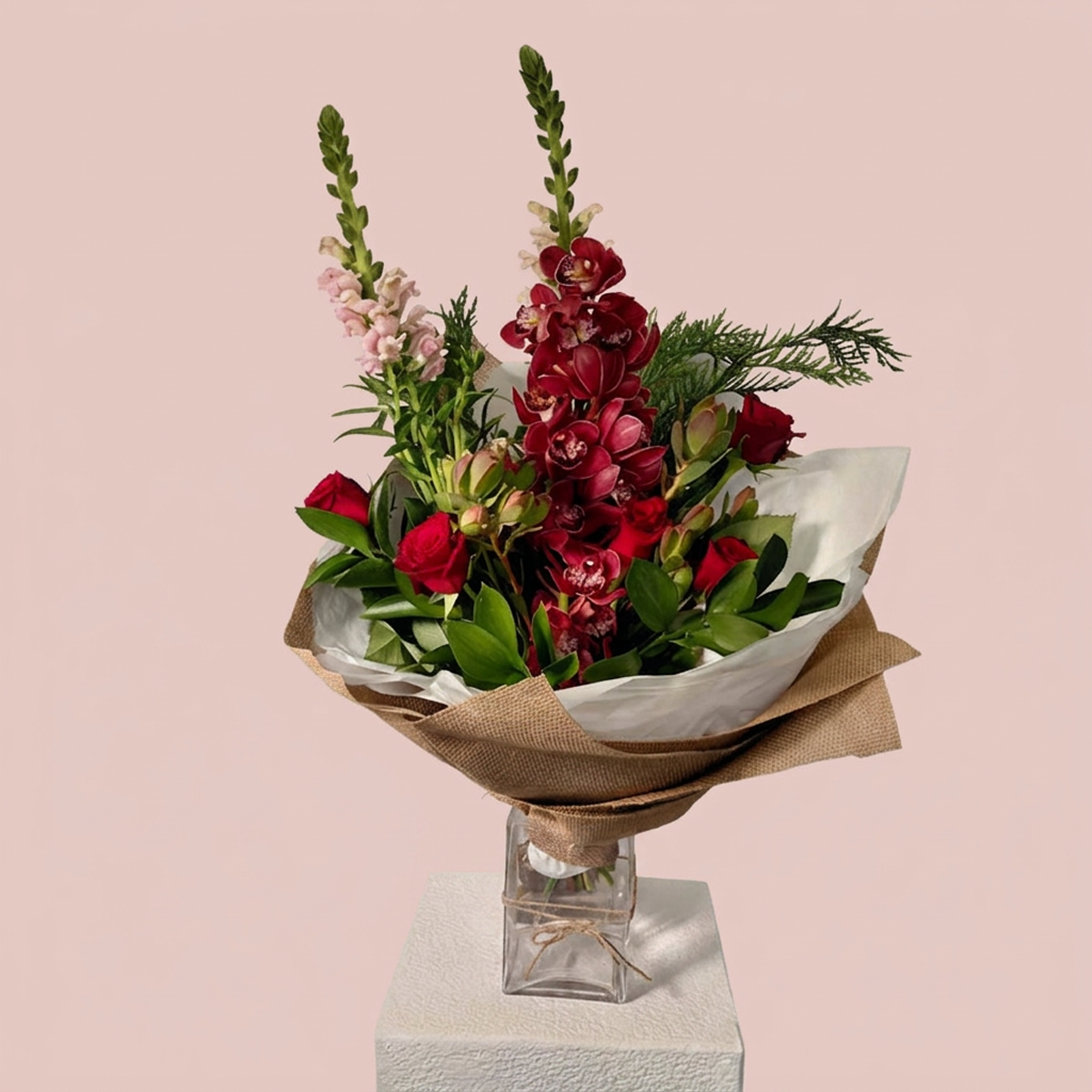 A floral bouquet with red flowers, green foliage, and light pink blossoms wrapped in brown paper and white tissue, displayed in a clear glass vase on a white pedestal against a soft pink background.