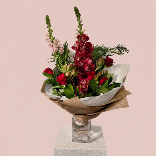 A floral bouquet with red flowers, green foliage, and light pink blossoms wrapped in brown paper and white tissue, displayed in a clear glass vase on a white pedestal against a soft pink background.