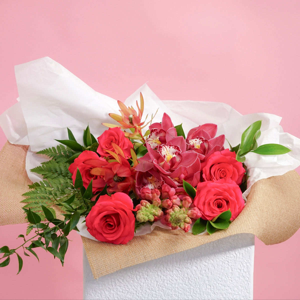 A vibrant floral bouquet featuring red roses, pink orchids, and green foliage, wrapped in white tissue paper and natural burlap, displayed on a white pedestal against a solid pink background.