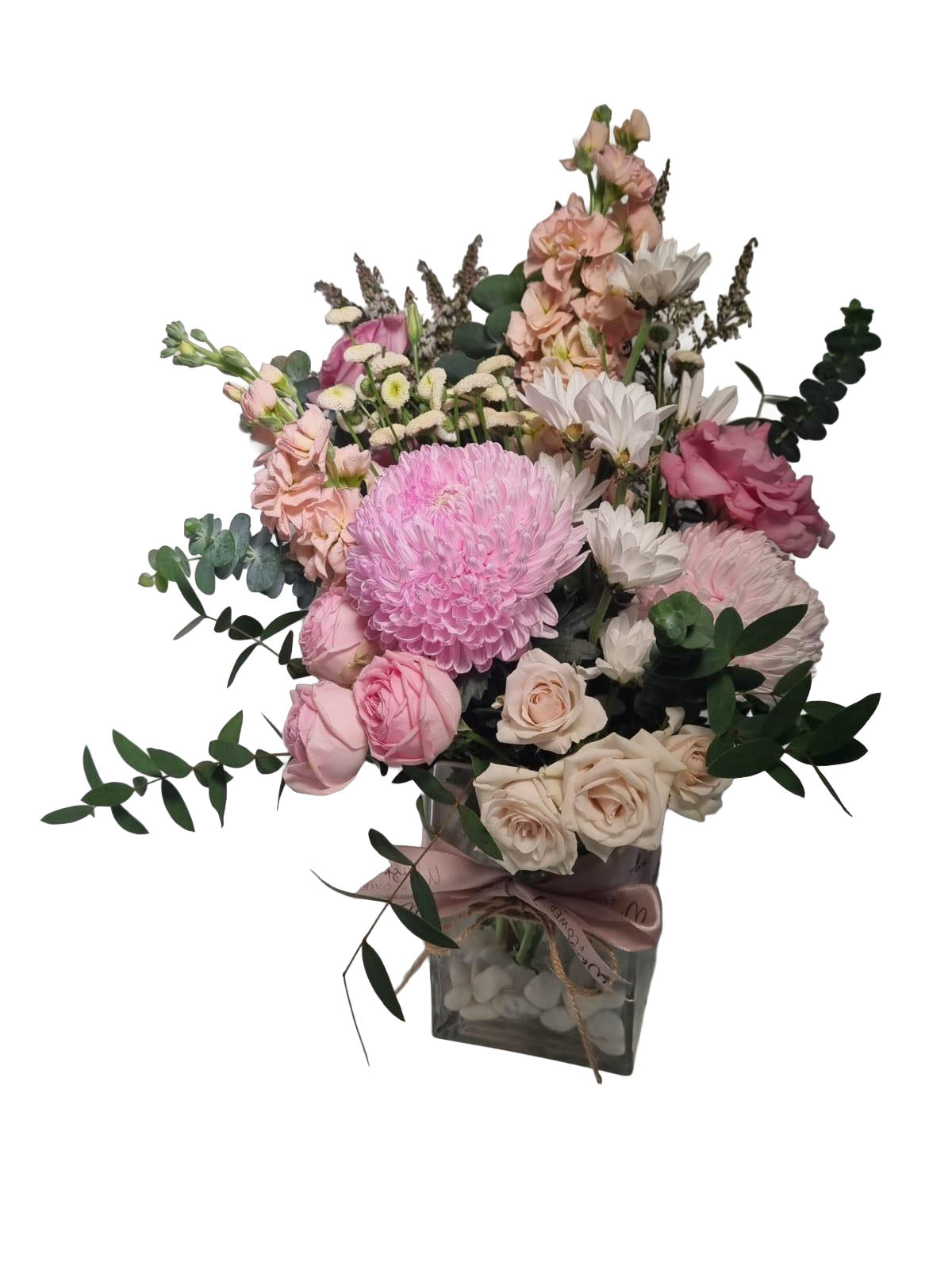 A square floral arrangement in a clear glass vase, featuring pink chrysanthemums, roses, carnations, and green foliage, tied with a dusty pink ribbon on a white background.