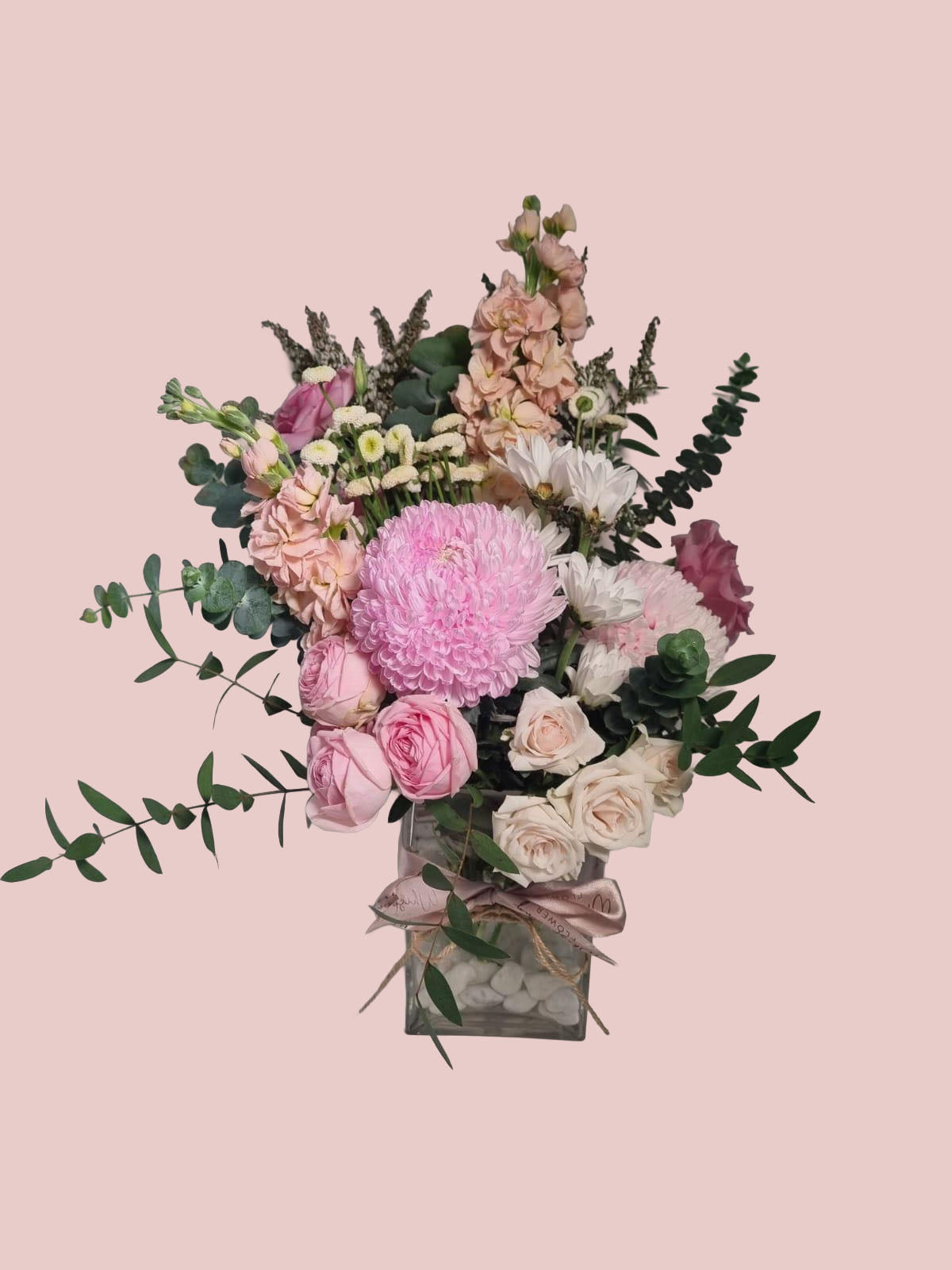 A floral arrangement featuring pink and white flowers with green foliage, presented in a square glass vase tied with a pink ribbon, set against a solid pale pink background.