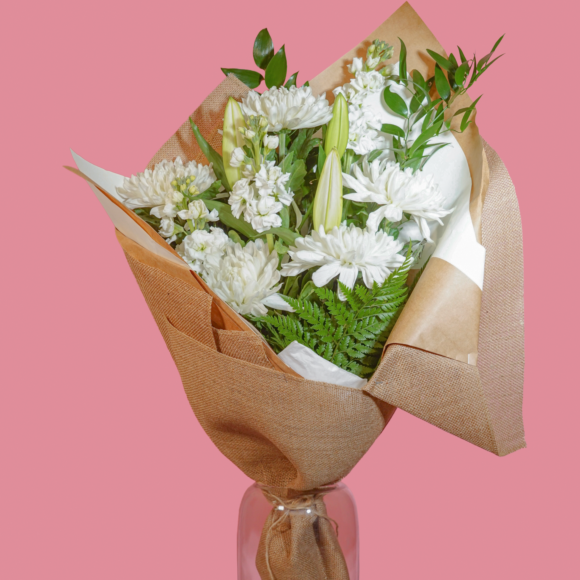 A bouquet of white flowers wrapped in brown kraft paper and displayed in a clear glass vase on a white pedestal against a solid pink background.