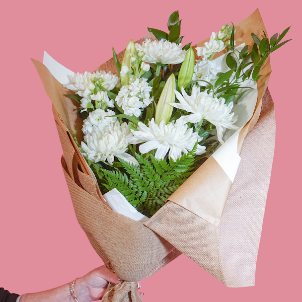 A bouquet of white flowers and green foliage wrapped in brown burlap and kraft paper, held by a hand against a solid pink background.