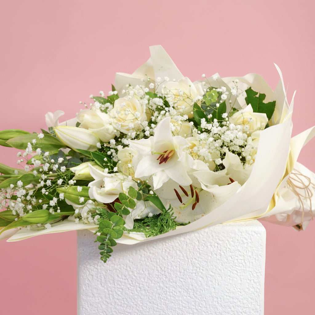 A bouquet of white lilies, white roses, and baby's breath flowers with green leaves, wrapped in white tissue paper and tied with a gold string, displayed on a white pedestal against a solid pink background.
