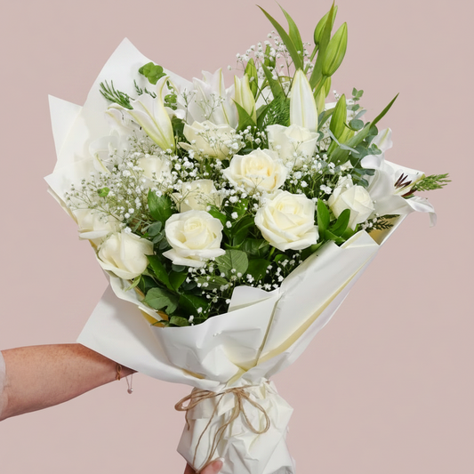 A bouquet of white roses, lilies, and baby's breath flowers wrapped in white paper and held by two hands against a solid pink background.