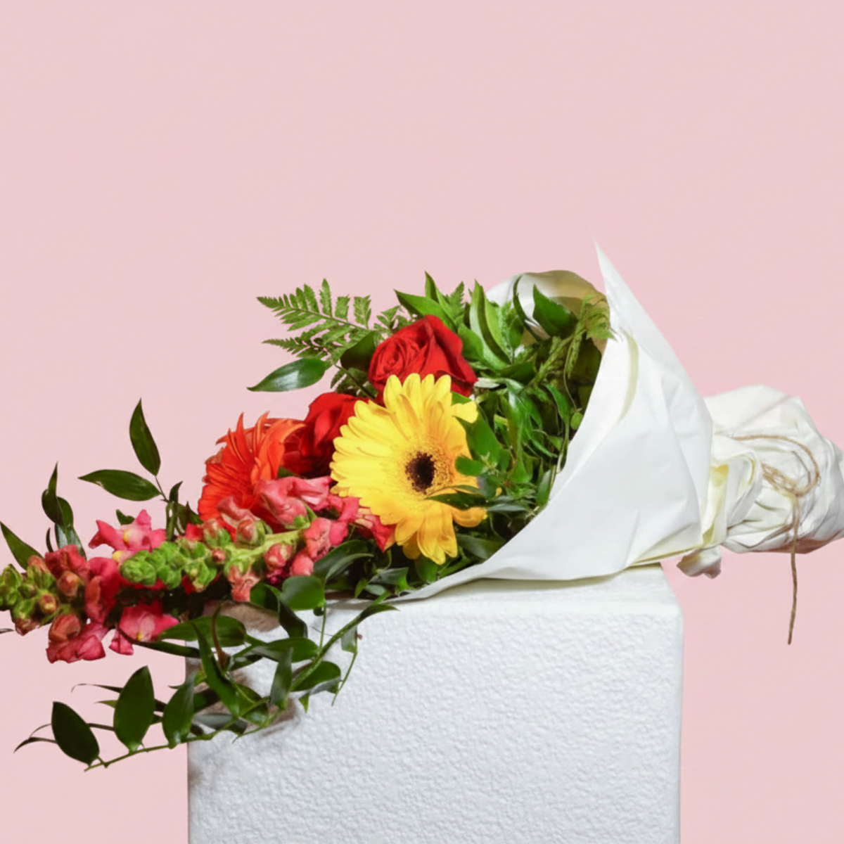 A vibrant floral bouquet wrapped in white paper, featuring a bright yellow gerbera daisy, red roses, pink snapdragons, and green foliage, displayed on a white textured pedestal against a solid pink background.