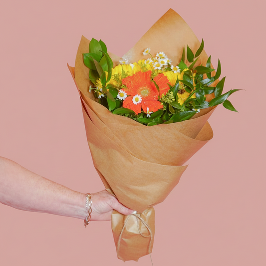 A hand wrapped in a gold bracelet holds a bouquet of colorful flowers, including a bright orange gerbera daisy, yellow flowers, and white daisies, wrapped in brown kraft paper, set against a solid pink background.