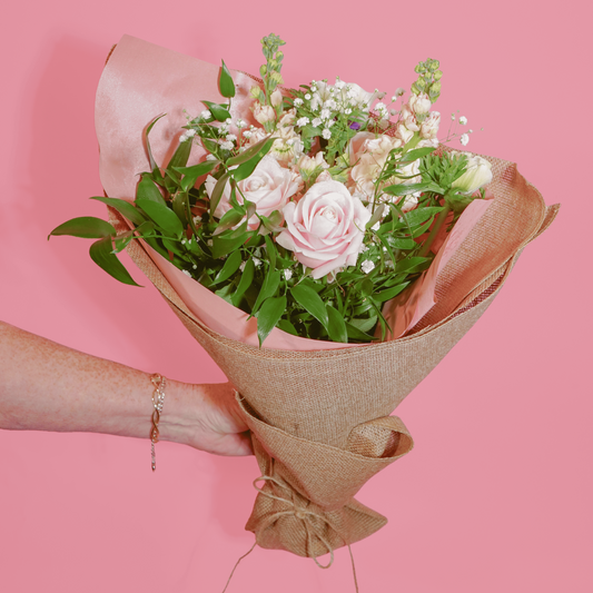 A hand holding a bouquet of pink roses, green foliage, and white baby's breath wrapped in pink tissue paper and brown burlap, set against a solid pink background.