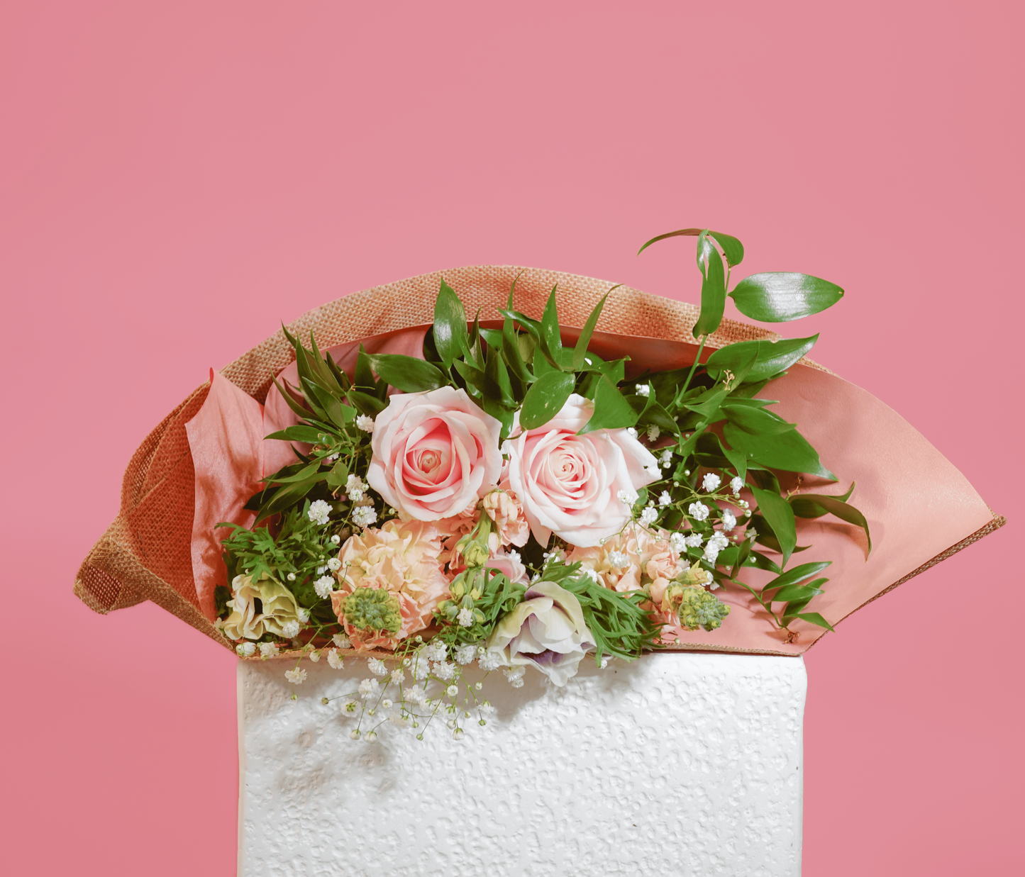 A floral arrangement featuring two pink roses, peach chrysanthemums, and green foliage, wrapped in peach paper and displayed on a white block against a solid pink background.