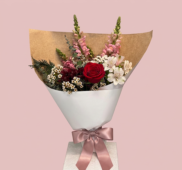 A floral bouquet wrapped in white paper with a dusty pink satin ribbon, featuring a red rose, pink flowers, white blossoms, and green foliage, set against a solid pink background.