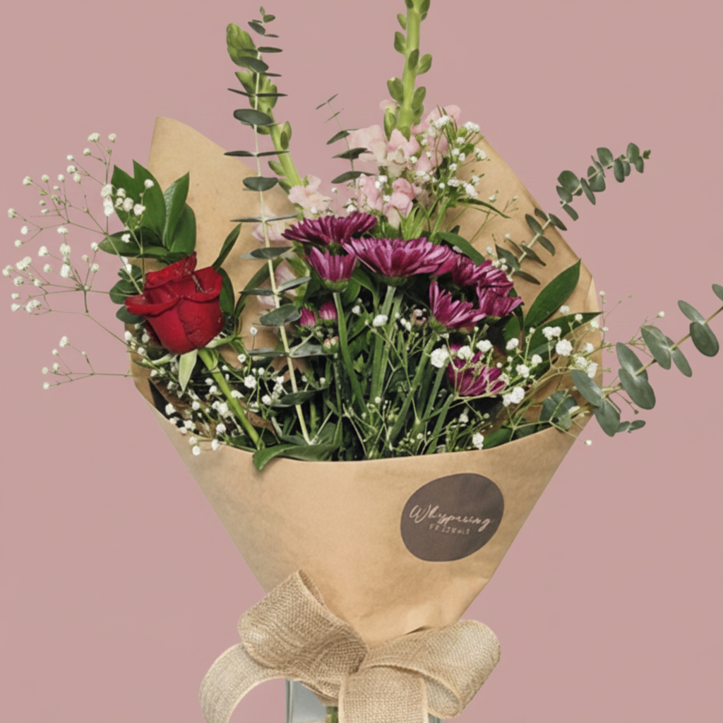 A fresh flower bouquet in a clear glass vase, wrapped in brown kraft paper with a beige ribbon bow. The bouquet features a red rose, purple daisies, pink flowers, green foliage, and white baby's breath, displayed on a white pedestal against a white background.