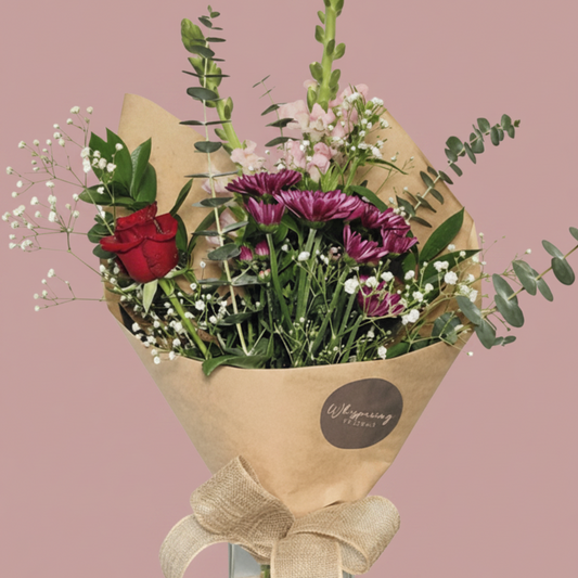 A fresh flower bouquet in a clear glass vase, wrapped in brown kraft paper with a beige ribbon bow. The bouquet features a red rose, purple daisies, pink flowers, green foliage, and white baby's breath, displayed on a white pedestal against a white background.