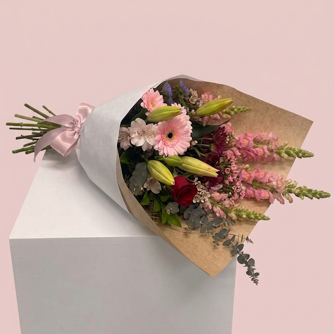 A wrapped flower bouquet with pink, red, white, and green flowers, tied with a pale pink ribbon and wrapped in brown kraft paper, displayed on a white cube against a soft pink background.