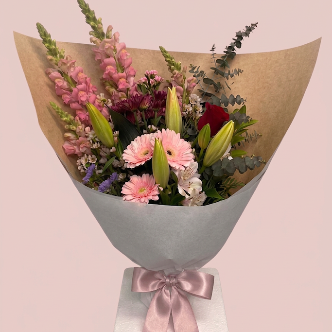 A wrapped flower bouquet featuring pink snapdragons, pink and red gerbera daisies, lily buds, burgundy roses, and green eucalyptus stems, wrapped in brown paper and light gray tissue with a pale pink satin ribbon, set against a solid pale pink background.