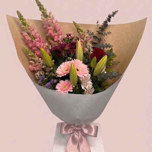 A wrapped flower bouquet featuring pink snapdragons, pink and red gerbera daisies, lily buds, burgundy roses, and green eucalyptus stems, wrapped in brown paper and light gray tissue with a pale pink satin ribbon, set against a solid pale pink background.