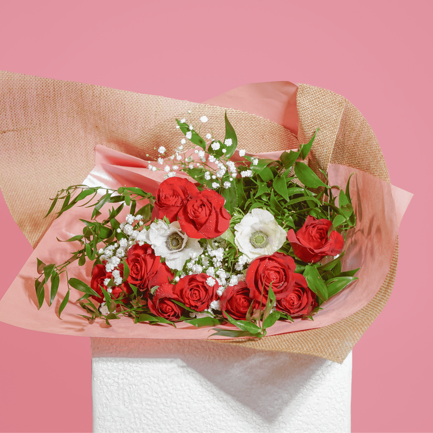 A bouquet of red roses, white flowers, and green foliage wrapped in pink and burlap-style paper, displayed on a white pedestal against a solid pink background.