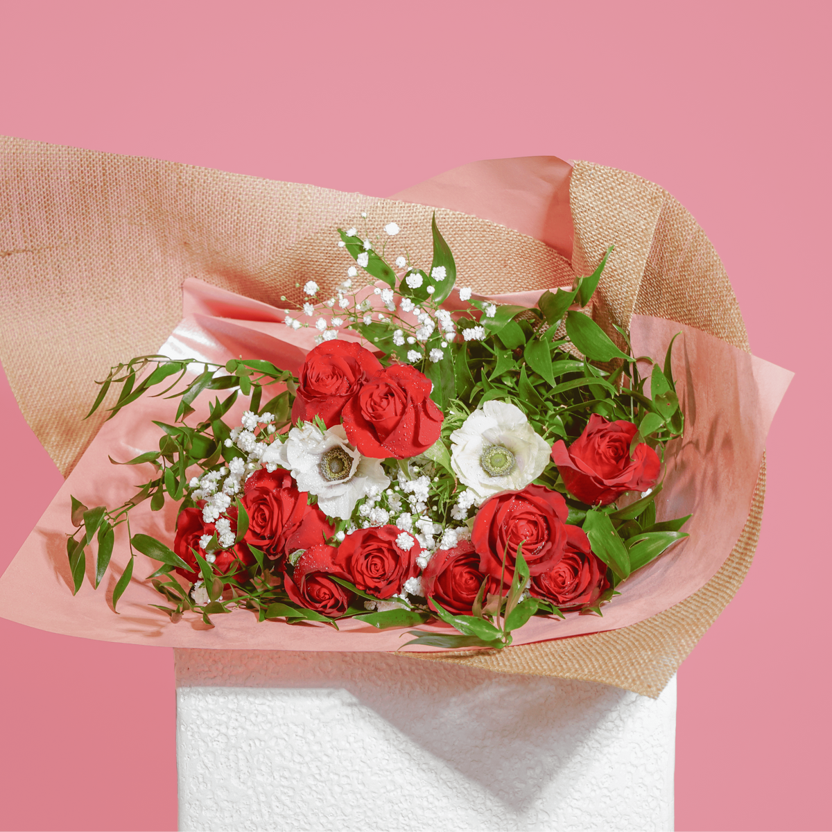 A bouquet of red roses, white flowers, and green foliage wrapped in pink and burlap-style paper, displayed on a white pedestal against a solid pink background.