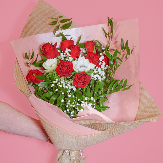 A bouquet of red and white roses with green foliage, wrapped in pink tissue paper and natural burlap, held by a person's hand against a solid pink background.