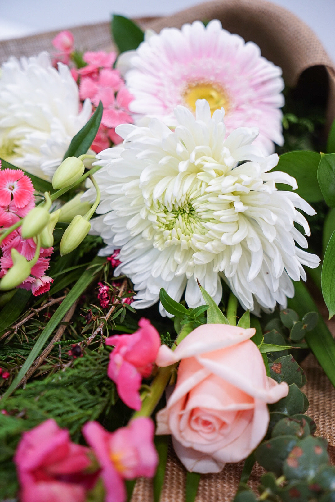 A close-up of a floral arrangement featuring white and light pink flowers, including a large white chrysanthemum, a pale pink rose, pink carnations, and green foliage, set against a burlap-like background.